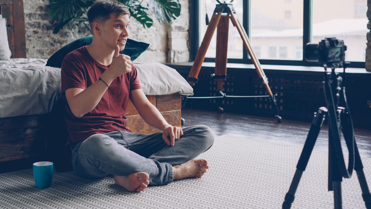 Young man sitting on the floor filming a video at home, surrounded by tripod and camera.